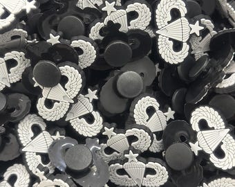 Peut inclure: Une image en gros plan d'un tas de breloques en caoutchouc noir et blanc en forme de badge de parachutiste. Les breloques ont une étoile blanche et un parachute blanc avec des ailes.