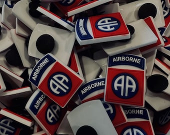 Peut inclure: Gros plan sur un tas d'aimants blancs et rouges avec le mot "Airborne" et un logo "AA" stylisé. Les aimants sont conçus pour être fixés aux chaussures ou aux sacs.