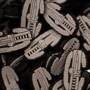 May include: A close-up of a pile of black and white military patches. The patches are shaped like a wreath with a knife in the center. The patches are made of a soft, rubbery material.