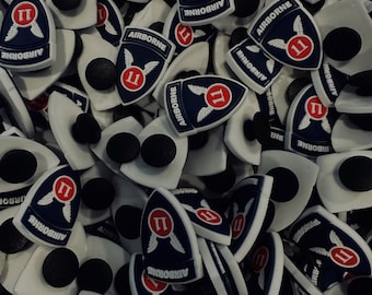 Peut inclure: Gros plan sur un tas de breloques pour chaussures blanches, bleues et rouges. Chaque breloque présente un bouclier bleu avec un chiffre romain rouge 'II' et le mot 'AIRBORNE' écrit en blanc. Les breloques sont conçues pour être attachées aux chaussures.