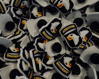 Peut inclure: Un grand groupe de breloques pour chaussures blanches, noires et jaunes représentant l'insigne de la 101e division aéroportée. Les breloques sont en caoutchouc et ont un design 3D. Les breloques représentent un aigle blanc aux ailes noires et au bec jaune, perché sur un bouclier noir avec le mot "AIRBORNE" écrit en lettres blanches.