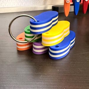 May include: A metal keyring holding a collection of five oval-shaped plastic tags. Each tag features a distinct solid color—blue, yellow, green, orange, and purple—layered with horizontal white stripes. Each tag has a small circular hole at one end.