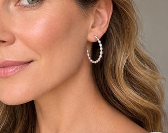 May include: A hoop earring featuring a continuous row of small, white, oval-shaped pearls. The pearls are arranged in a circular formation, creating a delicate and refined look. The earring is secured through the earlobe with a thin metal post.