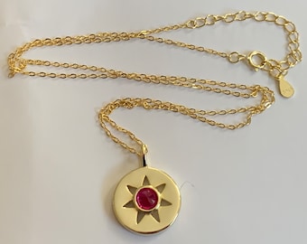 May include: A gold chain necklace with a pendant featuring a red gemstone set in a star-shaped cutout. The pendant is round and has a gold finish.