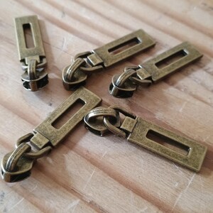 May include: Five antique brass-toned metal zipper pulls, each featuring a rectangular tab with a central cutout slot. These hardware components are designed for attachment to zipper sliders, showcasing a weathered, metallic finish throughout.