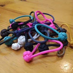 May include: A collection of zipper pulls made from paracord, each featuring a spherical monkey fist knot. The pulls are available in various colors including bright pink, turquoise, navy blue, white, black, olive green, and mustard yellow.