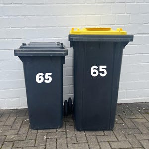 Könnte beinhalten: Zwei schwarze Mülltonnen mit gelben Deckeln. Die größere Tonne hat die Nummer 65 auf der Vorderseite. Die kleinere Tonne hat die Nummer 65 auf der Vorderseite.