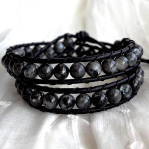 May include: A wrap bracelet featuring dark gray, round gemstone beads. The beads are strung on multiple strands of black leather. The bracelet is tied with a knot closure. The background is a white, textured fabric.
