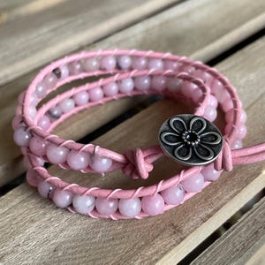 May include: A wrap bracelet featuring pink beads and pink leather. The bracelet wraps around the wrist multiple times and is secured with a silver-colored flower-shaped button clasp. The beads are a light pink color.