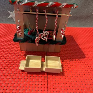 May include: A miniature wooden Christmas market stall with a red and green striped roof, decorated with candy canes and a heart-shaped gingerbread cookie. The stall has two wooden boxes in front of it.