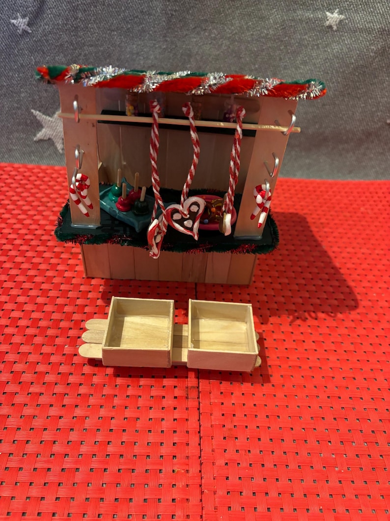 May include: A miniature wooden Christmas market stall with a red and green striped roof, decorated with candy canes and a heart-shaped gingerbread cookie. The stall has two wooden boxes in front of it.