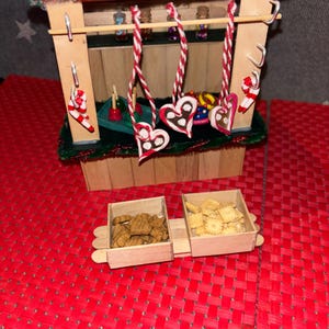 May include: A miniature wooden Christmas market stall with a red and green striped roof. The stall has a shelf with small jars of candy and a counter with gingerbread hearts and candy canes hanging from the ceiling. There are two wooden boxes filled with cookies in front of the stall.