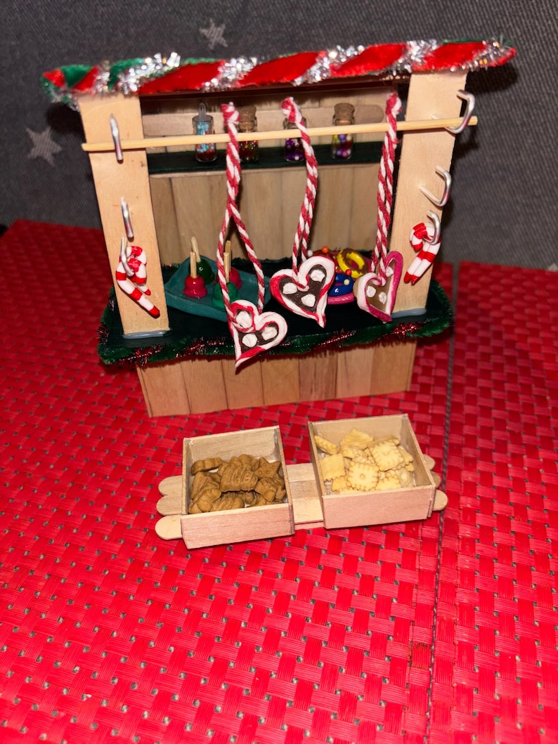 May include: A miniature wooden Christmas market stall with a red and green striped roof. The stall has a shelf with small jars of candy and a counter with gingerbread hearts and candy canes hanging from the ceiling. There are two wooden boxes filled with cookies in front of the stall.