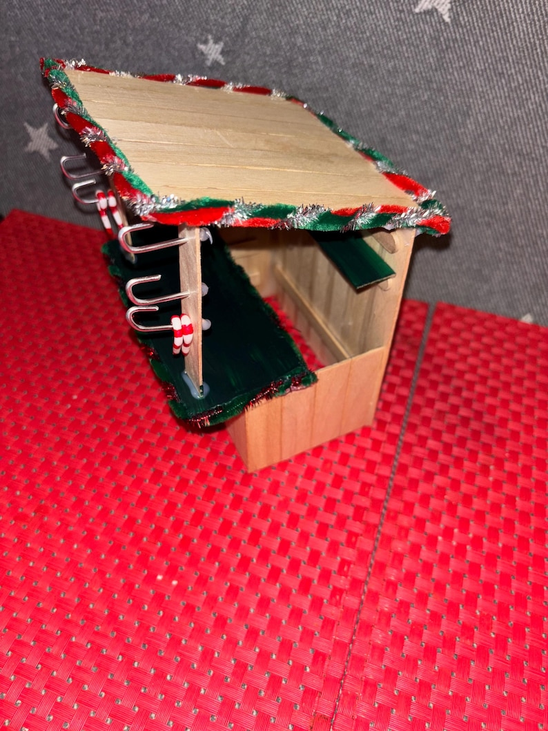 May include: A small wooden Christmas market stall with a green roof and red and white candy cane trim. The stall has hooks on the side for hanging items.