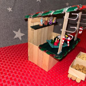 May include: A miniature wooden Christmas market stall with a green roof, candy cane decorations, and a variety of colorful candies in jars. The stall also has a green felt counter with candy apples and a wooden box filled with cookies.