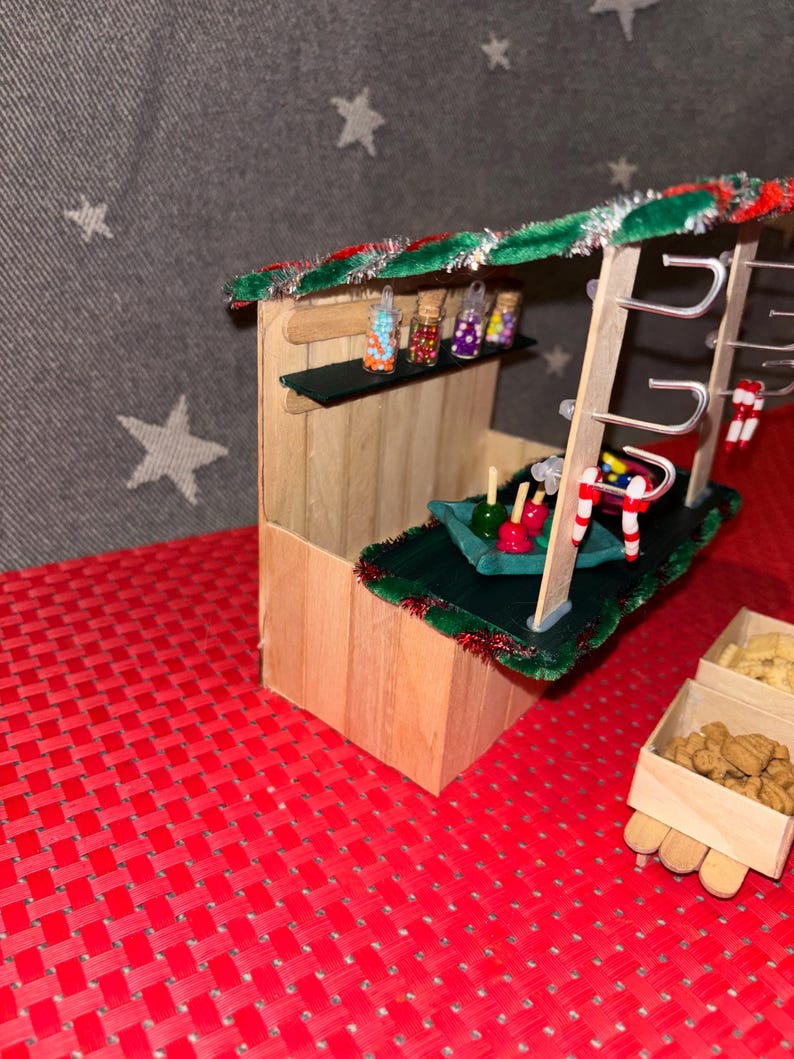 May include: A miniature wooden Christmas market stall with a green roof, candy cane decorations, and a variety of colorful candies in jars. The stall also has a green felt counter with candy apples and a wooden box filled with cookies.