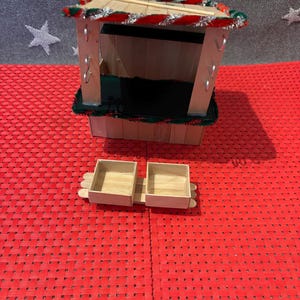 May include: A miniature wooden Christmas market stall with a red and green striped roof, a black base, and two small wooden boxes.