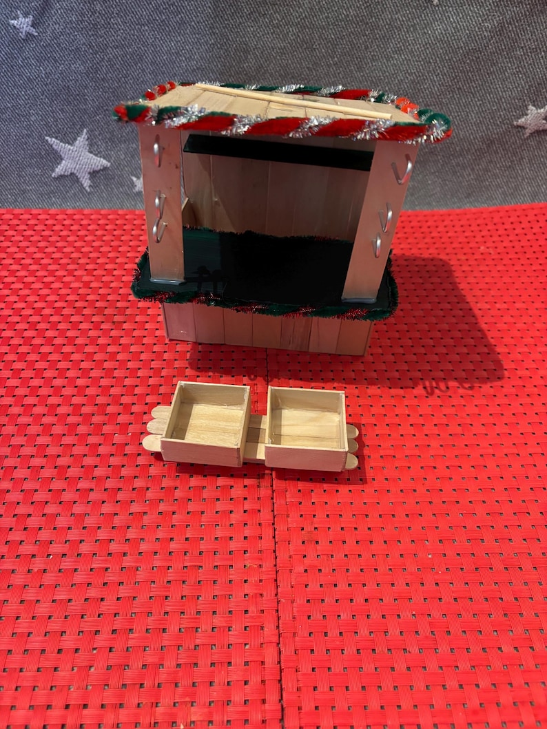 May include: A miniature wooden Christmas market stall with a red and green striped roof, a black base, and two small wooden boxes.