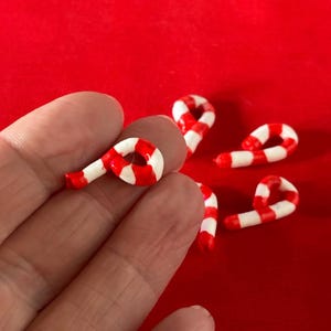 以下が含まれることがあります： 赤と白のキャンディーケーンの形をしたオーナメントが5つ。オーナメントは小さく、赤い表面の上に置かれています。