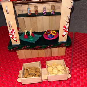 May include: A miniature wooden Christmas market stall with a green roof and red trim. The stall has a shelf with four small jars filled with colorful candies. There are also three wooden trays with various treats, including candy apples, donuts, and cookies.