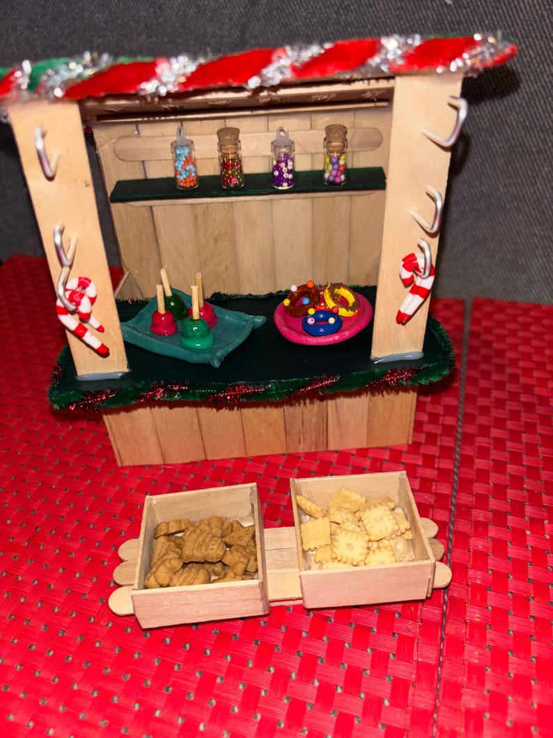 May include: A miniature wooden Christmas market stall with a green roof and red trim. The stall has a shelf with four small jars filled with colorful candies. There are also three wooden trays with various treats, including candy apples, donuts, and cookies.