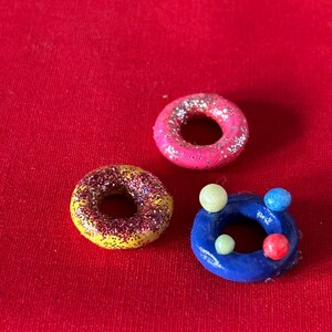 以下が含まれることがあります： ピンク、イエロー、ブルーの小さなキラキラしたドーナツが3つ。青いドーナツには小さな白と赤のボールが付いています。
