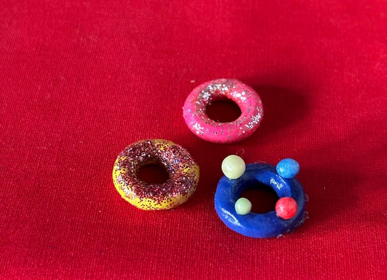 以下が含まれることがあります： ピンク、イエロー、ブルーの小さなキラキラしたドーナツが3つ。青いドーナツには小さな白と赤のボールが付いています。