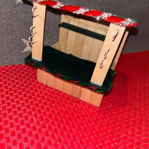 May include: A small wooden Christmas market stall with a red and green striped roof, a green counter, and silver hooks on the side. The stall is sitting on a red and white checkered surface.