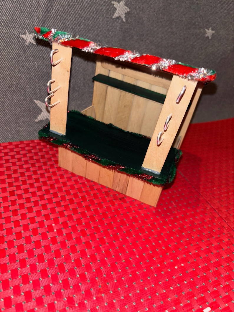May include: A small wooden Christmas market stall with a red and green striped roof, a green counter, and silver hooks on the side. The stall is sitting on a red and white checkered surface.