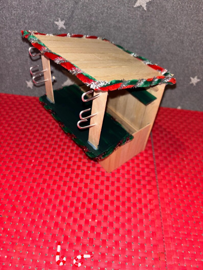 May include: A small wooden Christmas market stall with a green and red candy cane trim. The stall has hooks on the sides and a green floor.