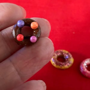 以下が含まれることがあります： カラフルなスプリンクルがトッピングされたチョコレートドーナツ、ピンクとパープルのスプリンクルがトッピングされたイエロードーナツ、白いスプリンクルがトッピングされたピンクドーナツの3つのミニドーナツ