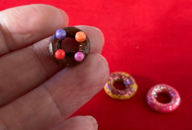 以下が含まれることがあります： カラフルなスプリンクルがトッピングされたチョコレートドーナツ、ピンクとパープルのスプリンクルがトッピングされたイエロードーナツ、白いスプリンクルがトッピングされたピンクドーナツの3つのミニドーナツ