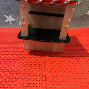 May include: A miniature wooden Christmas market stall with a red and green striped roof, a black counter, and silver hooks on the side.