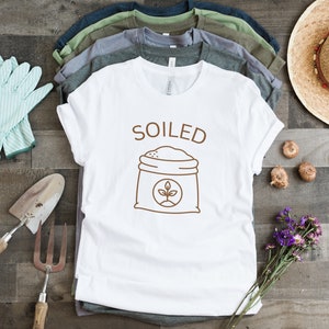 K&ouml;nnte beinhalten: Wei&szlig;es T-Shirt mit braunem Text, der "SOILED" sagt, und einer braunen Strichzeichnung eines Sackes Erde mit einer Pflanze, die daraus w&auml;chst.