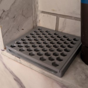 May include: A gray plastic shower drain with a honeycomb design. The drain is sitting on a white tiled surface.