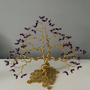 May include: A handcrafted wire tree sculpture with a gold-colored metal frame. The tree features branches adorned with purple and black beads, resembling blossoms. The base of the tree has a decorative, swirling design.