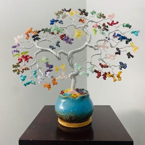 May include: A handcrafted wire tree sculpture with colorful gemstone leaves. The silver-toned wire branches are adorned with a variety of gemstones in shades of red, blue, green, yellow, and purple. The tree sits in a blue ceramic pot on a wooden base.