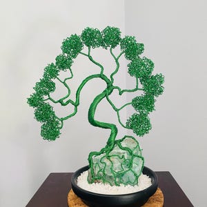 May include: A handcrafted wire tree sculpture with a green wire canopy and trunk, rooted in a light green stone base. The tree sits in a black ceramic pot filled with white pebbles, resting on a cork coaster. A decorative bonsai-style art piece.