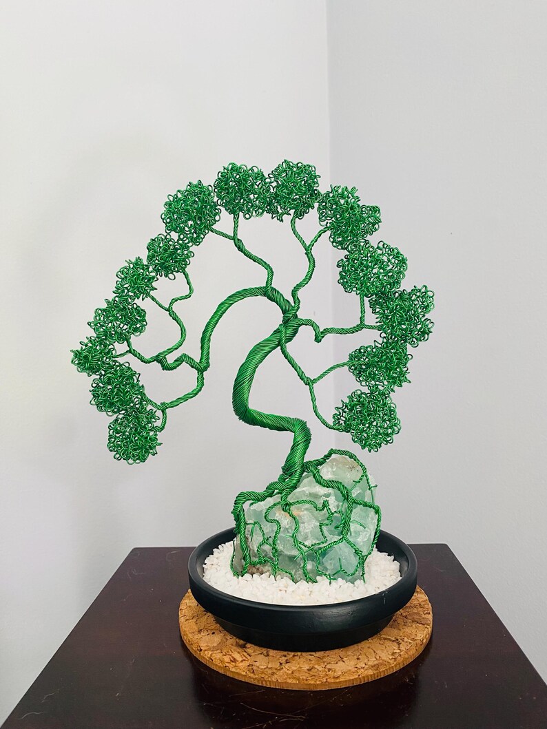 May include: A handcrafted wire tree sculpture with a green wire canopy and trunk, rooted in a light green stone base. The tree sits in a black ceramic pot filled with white pebbles, resting on a cork coaster. A decorative bonsai-style art piece.