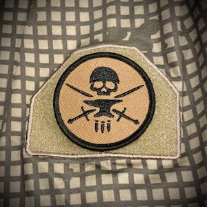May include: A tan and black embroidered patch featuring a skull and crossbones design. The skull is wearing a helmet and holding a hammer and anvil. The crossbones are made of swords. The patch has the Roman numeral III in the centre.