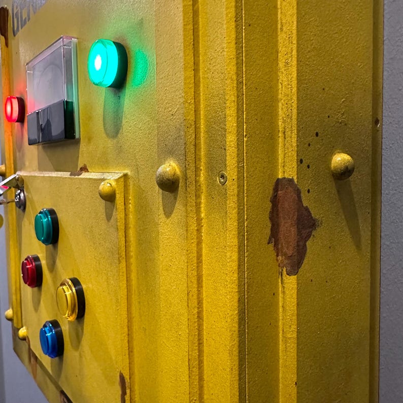 May include: A yellow metal control panel with a variety of buttons and a keyhole. The panel has a worn, distressed look with some rust spots. There are red, green, blue, and yellow buttons.