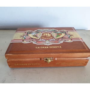 May include: A wooden cigar box with a hinged lid and brass clasp. The box is brown with a decorative label on the top and the words "LA GRAN OFERTA" printed on the front. The box is closed.