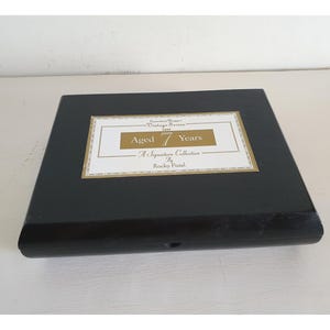 May include: A black wooden box with a rectangular label in the center. The label has a gold border and text that reads "Aged 7 Years" and "A Signature Collection By Rocky Patel."