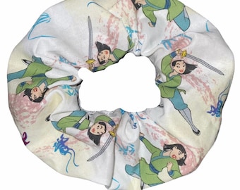 May include: White scrunchie with a repeating pattern of Mulan, the Disney princess, holding a sword. The background is a light pink and blue watercolor design.