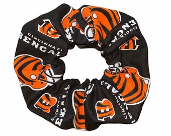 May include: Black and orange scrunchie with a repeating pattern of the Cincinnati Bengals logo. The logo features a tiger head with stripes and the words "Cincinnati Bengals".