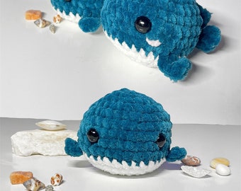 May include: Two teal blue crocheted whale plushies with white teeth and black eyes. The whales are sitting on a white surface with seashells.
