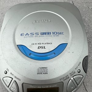 May include: A silver Aiwa CD player with a blue display screen. The player has buttons for skip, search, play, pause, stop, and open. The text on the player reads "E.A.S.S PLUS 10SEC ELECTRONIC ANTI SHOCK SYSTEM 1bit DAC CD-R/RW PLAYBACK DSL COMPACT DISC DIGITAL AUDIO COMPACT DISC PLAYER XP-V416C TRACK/TIME"
