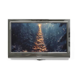 May include: A silver business card holder featuring a winter scene with a decorated Christmas tree. The artwork shows a snow-covered forest with a glowing tree, lit with warm lights, under a starry sky.