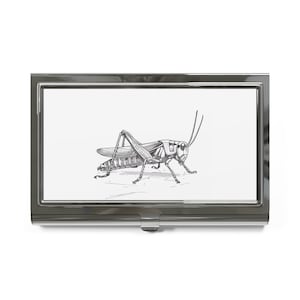 May include: A silver metal business card holder with a black and white line drawing of a grasshopper on a white background.