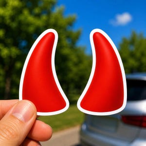 May include: Two separate red devil horn stickers with a white border. Each horn has a curved, tapered shape with a smooth, solid red surface. The stickers are presented as individual die-cut decals with a clean white outline around the edges.